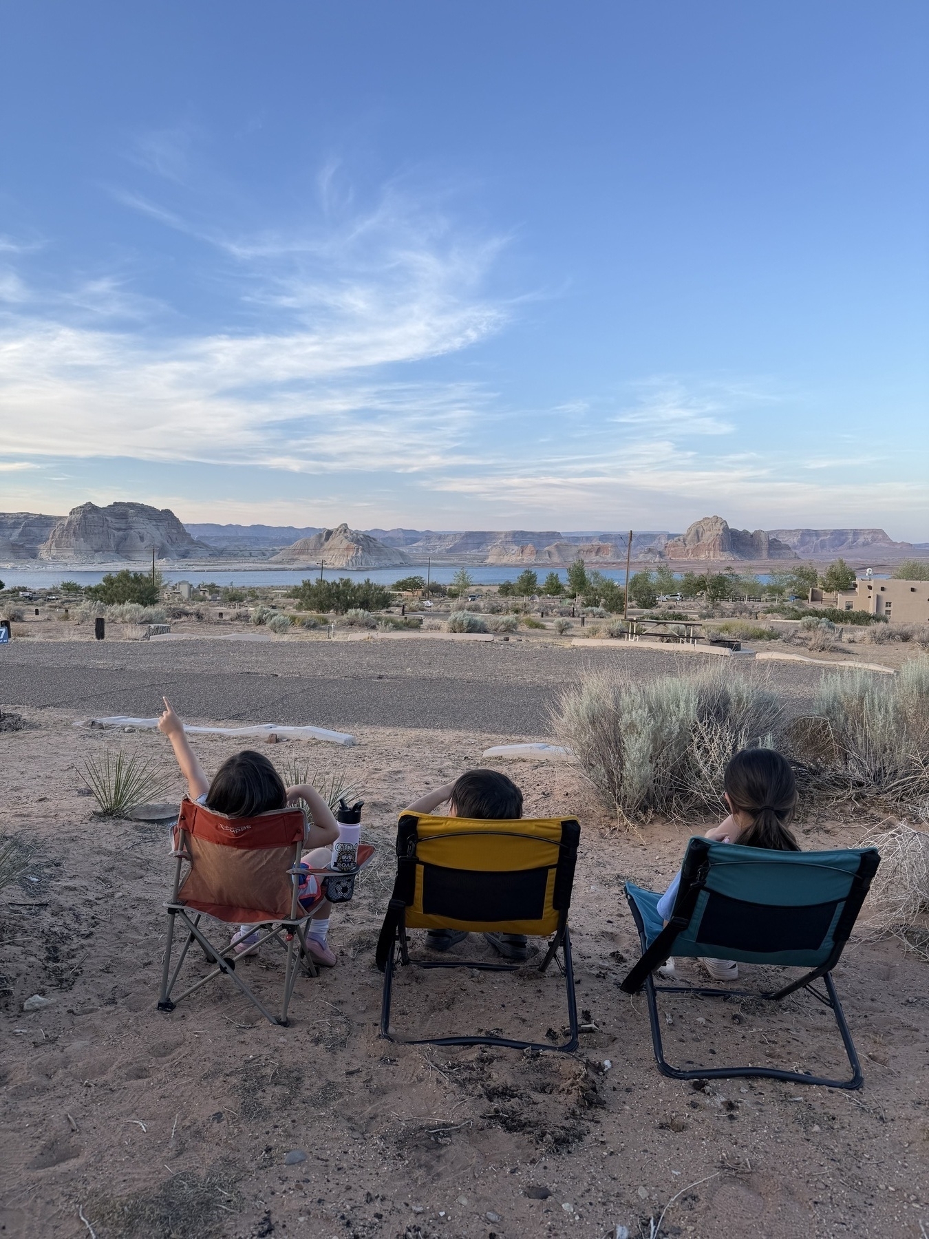 Three people are sitting in camping chairs, enjoying a scenic view of a desert landscape with distant rock formations under a partly cloudy sky.