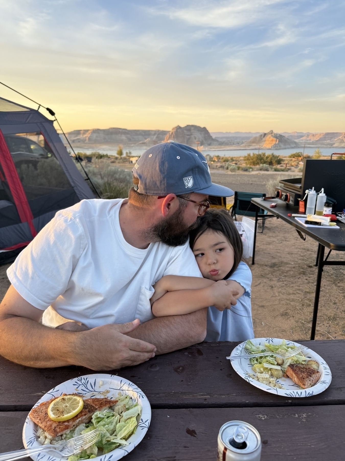 A man and a child, seated at a picnic table with plates of food, enjoy a campsite in a scenic desert landscape.