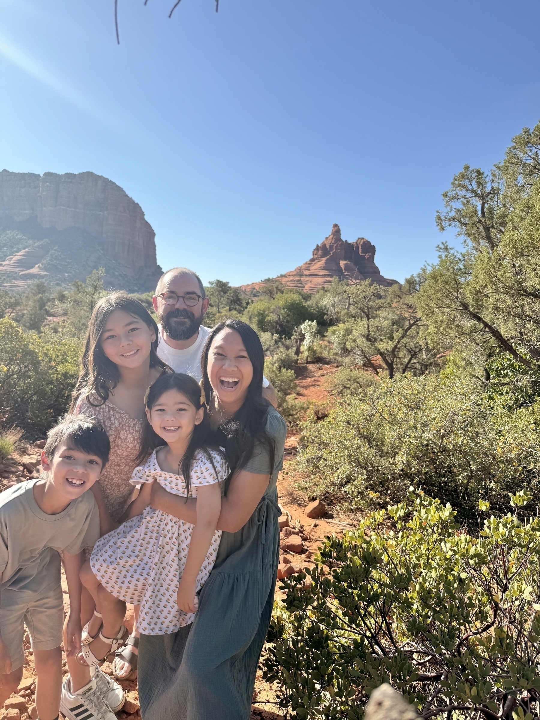 A family of five is smiling together in a sunny desert landscape with red rock formations in the background.