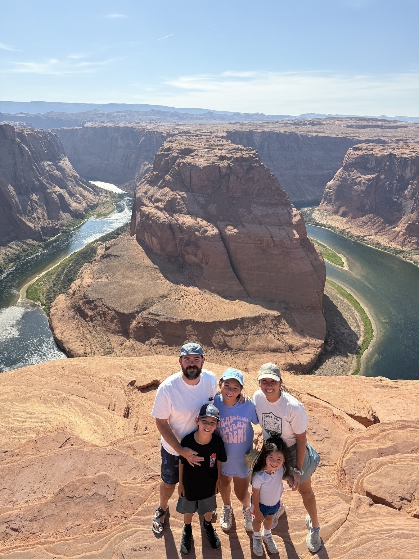 A family of five stands smiling on a rocky overlook with Horseshoe Bend and the Colorado River in the background under a clear blue sky.