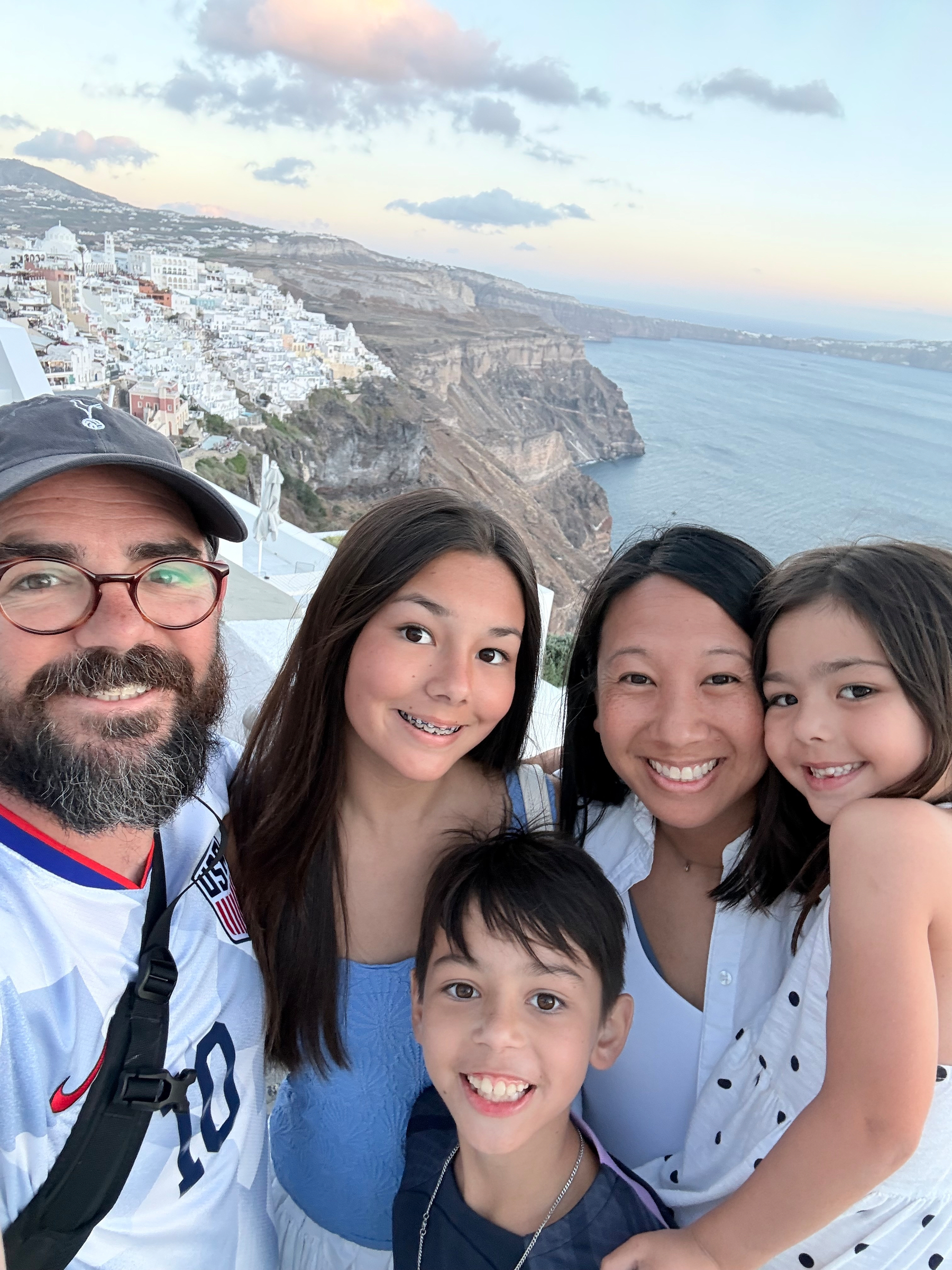 A family of five is smiling and posing for a selfie with a scenic coastal cliffside and white buildings in the background during sunset.